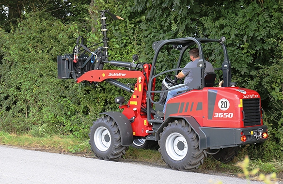 Schäffer 3650 loader trimming overgrown roadside hedges using a hydraulic hedge trimmer attachment.