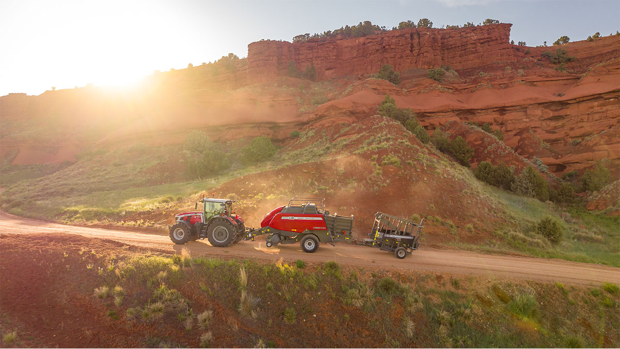 Hesston by Massey Ferguson LB2200 Series baler attached to Massey Ferguson tractor on red dirt road at sunrise in rugged terrain.