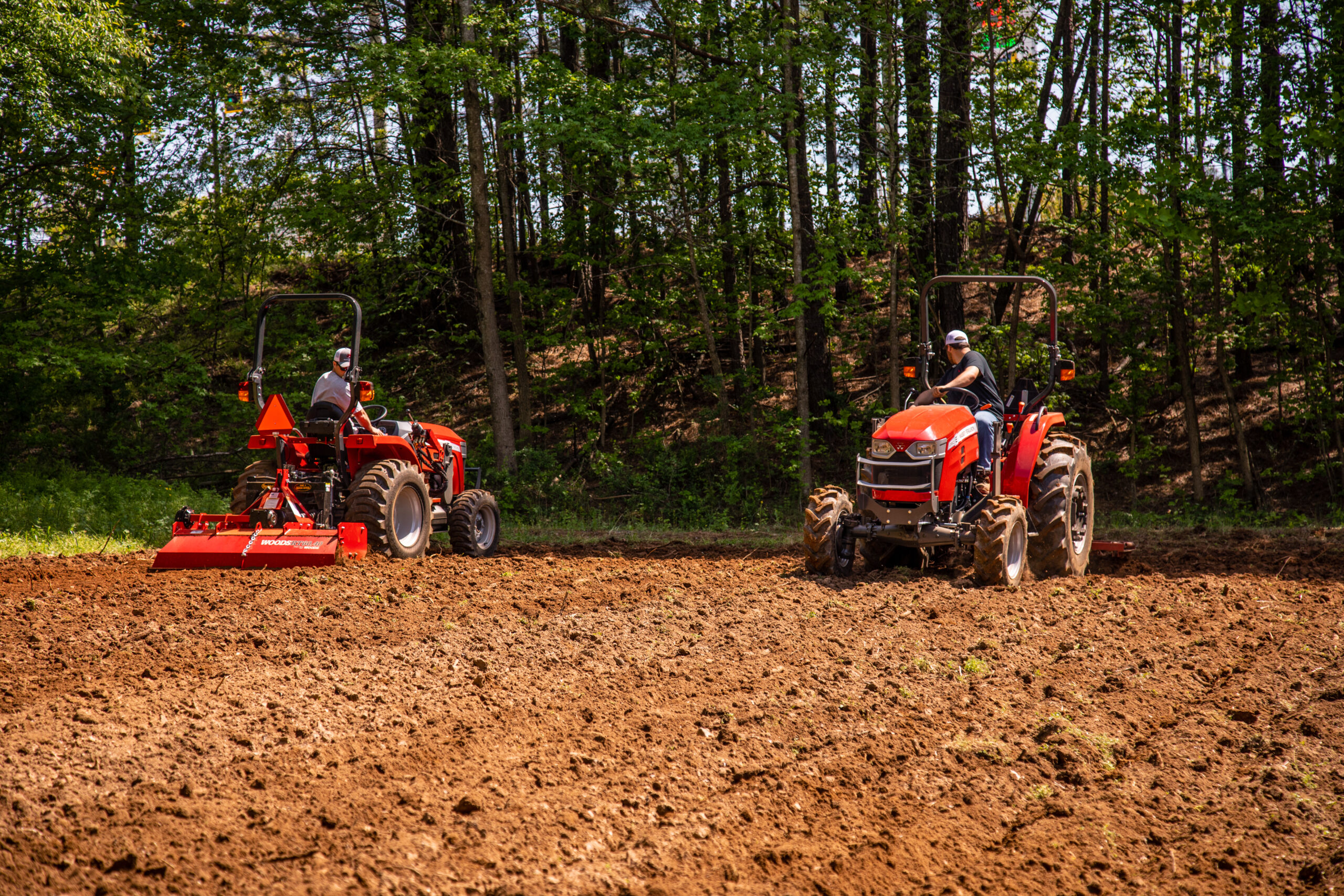 Two Massey Ferguson compact tractors tilling soil in a wooded field, showcasing MF 1800E and 2800E Series performance.