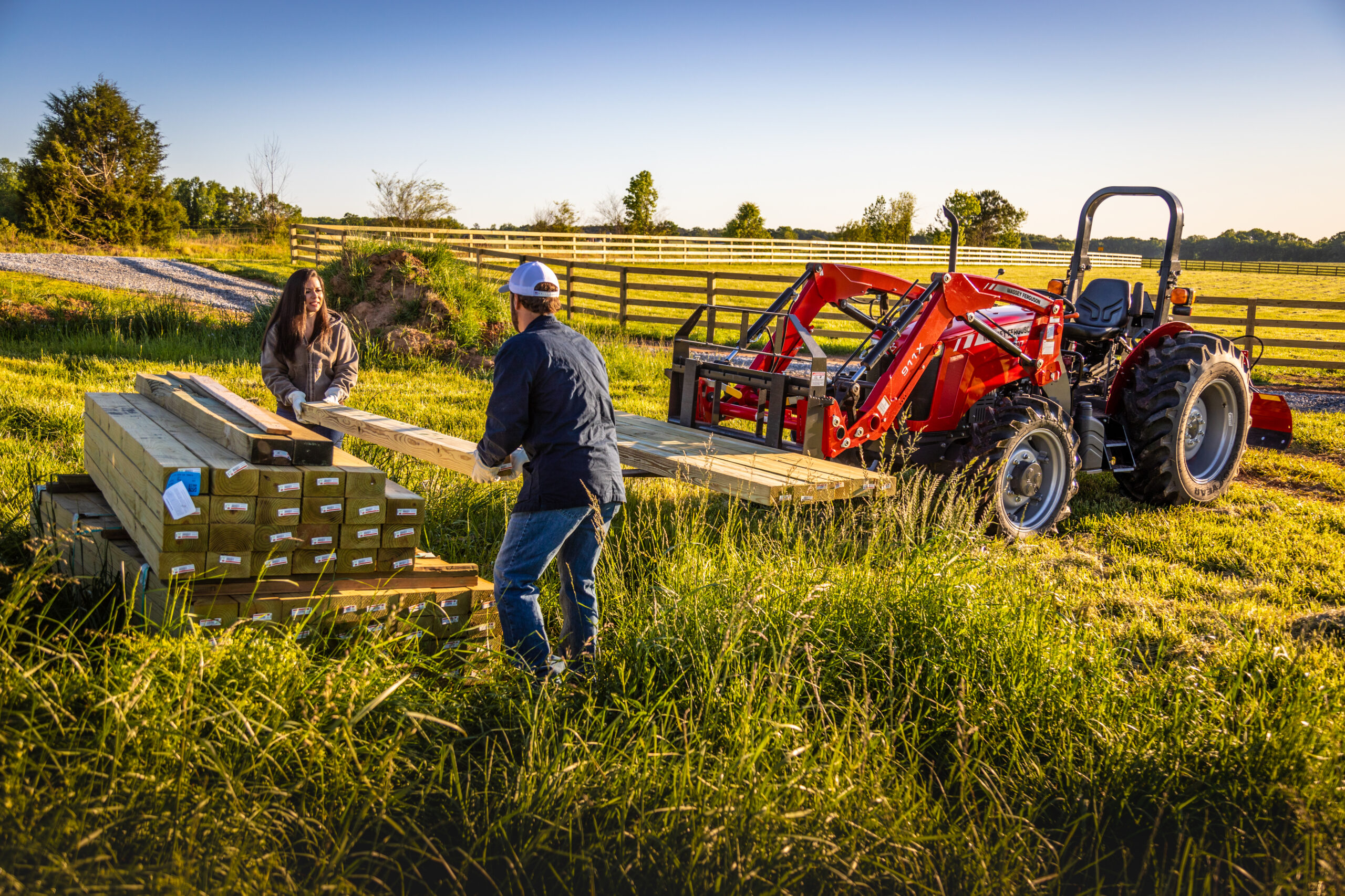 Two people loading lumber beside a Massey Ferguson 2600H Series utility tractor with loader, ideal for fencing and property maintenance.