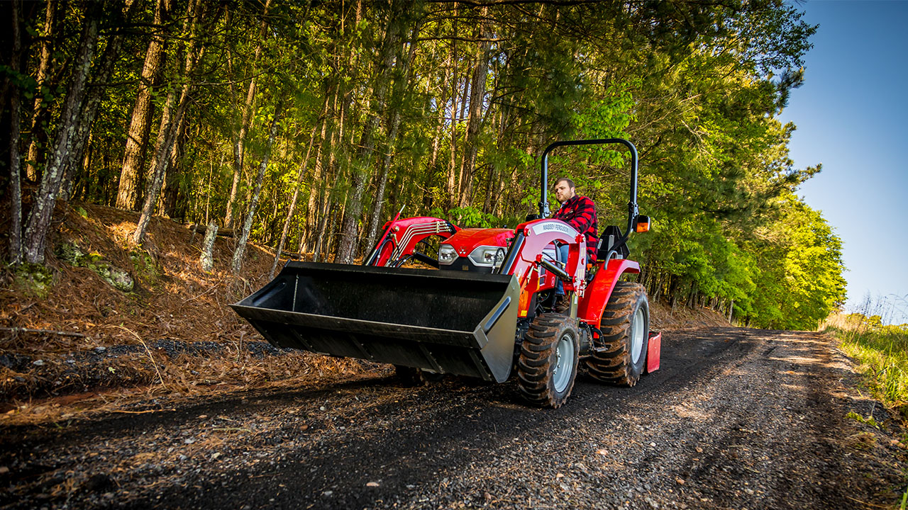 Massey Ferguson compact tractor driving on forest path with front loader raised, operated by man in red flannel shirt.