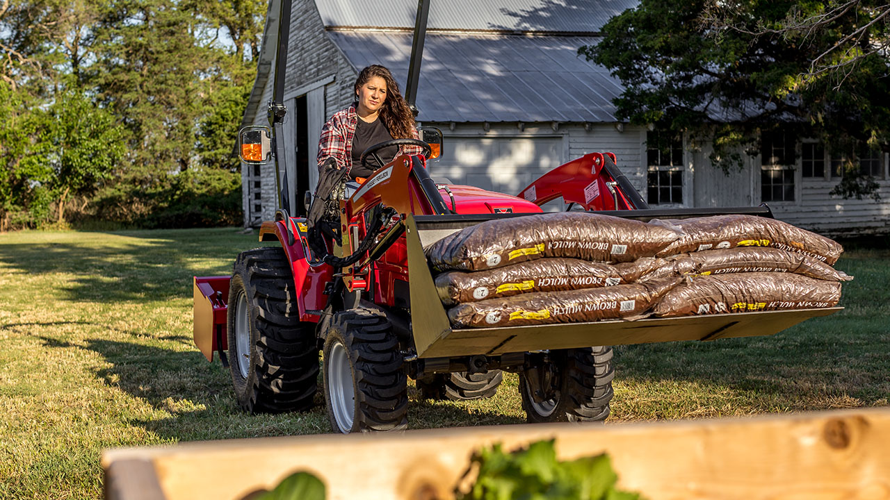 Woman driving Massey Ferguson 1526 tractor hauling bags of brown mulch in front loader on a residential property.