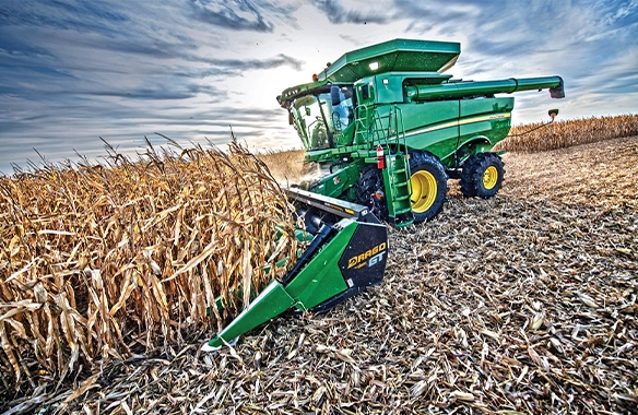John Deere combine harvesting corn with a Drago GT Series corn head, featuring automatic self-adjusting deck plates for reduced kernel loss.