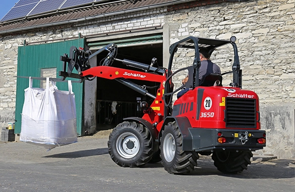 Schäffer 3650 transporting a large bulk feed bag using a bag lifter attachment in front of a livestock barn.