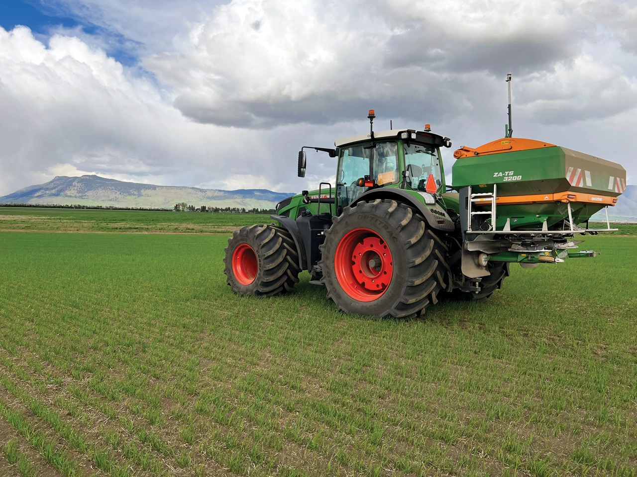 Fendt tractor equipped with an Amazone ZA-TS 3200 fertilizer spreader operating in a green field with mountains in the background under a cloudy sky.