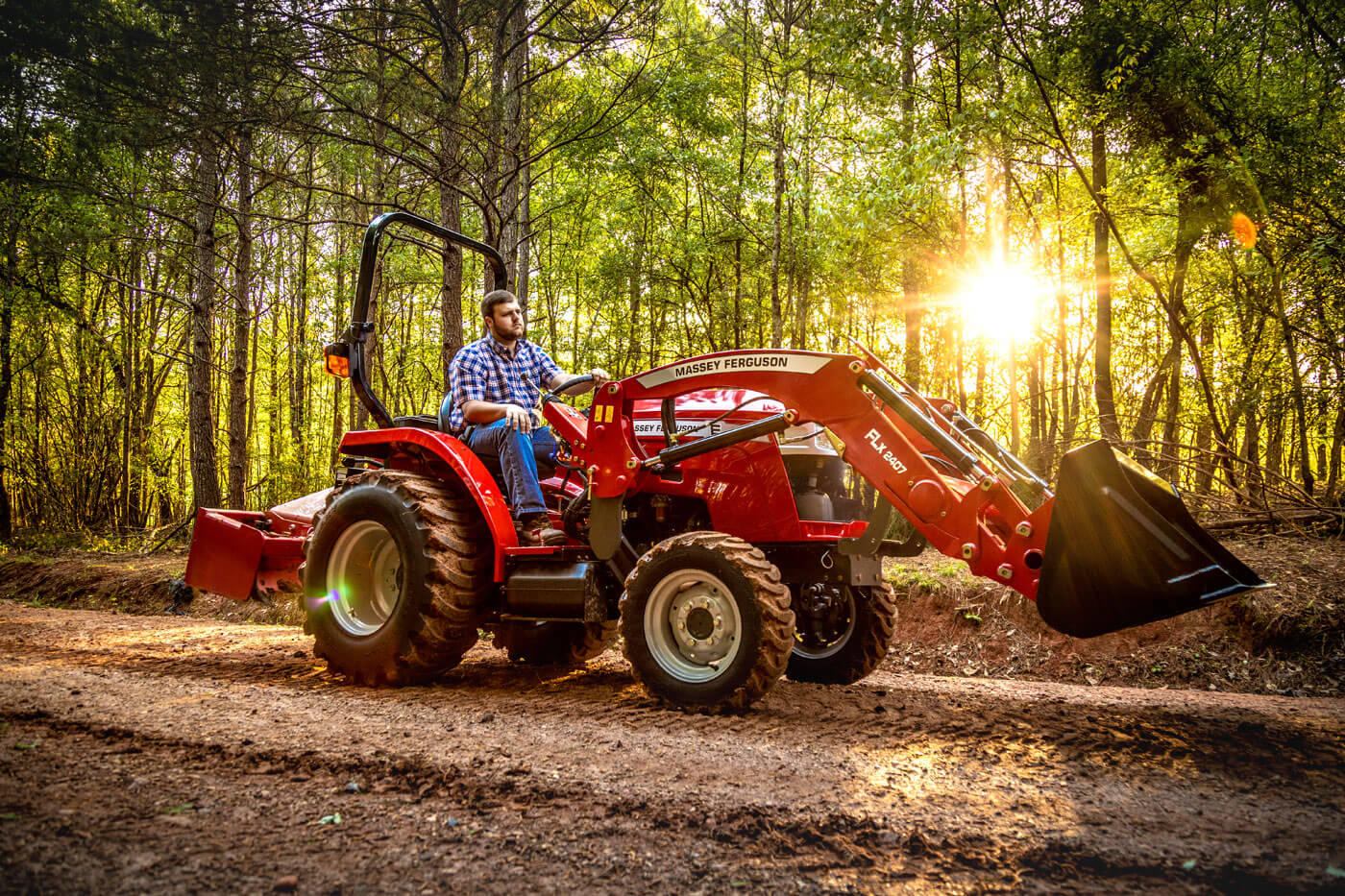 Man operating a Massey Ferguson 1800E compact tractor with front loader in a wooded area at sunset.