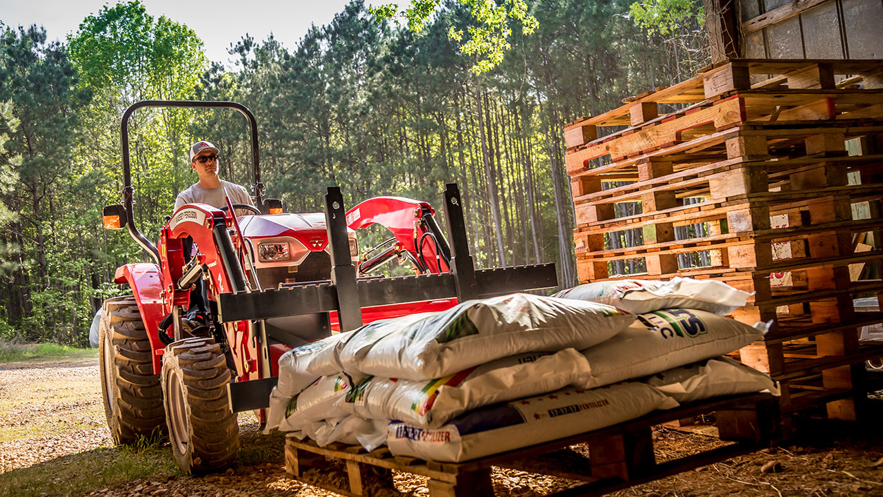 Operator using a Massey Ferguson compact tractor with pallet forks to move fertilizer bags near stacked wooden pallets.