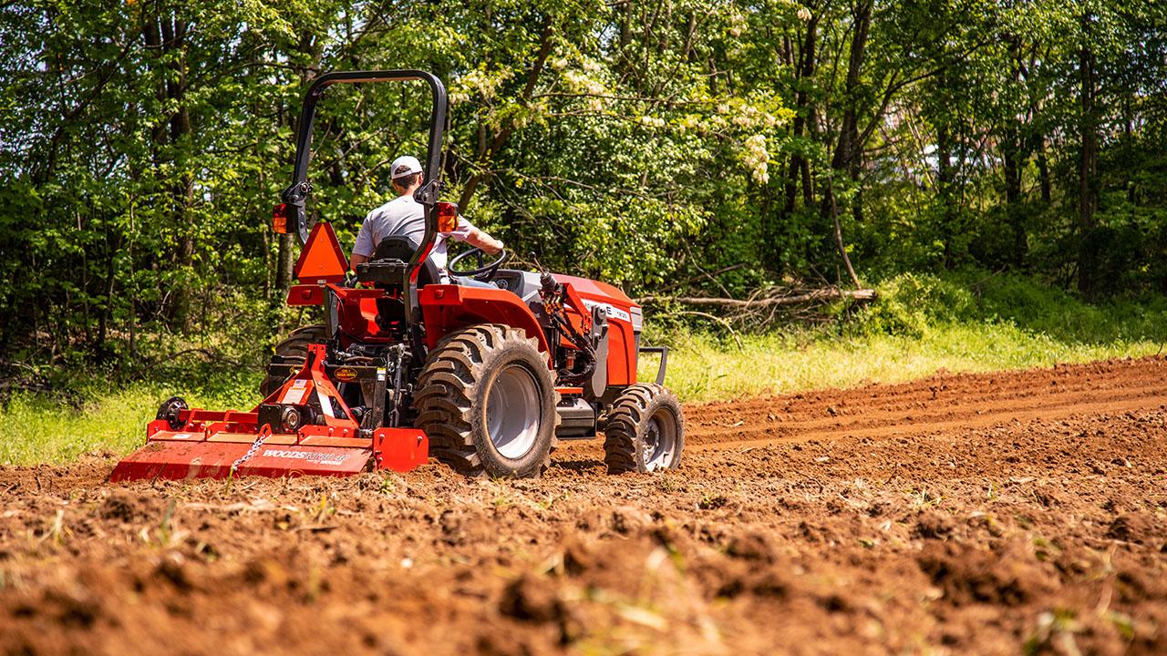Massey Ferguson 1800E tractor with Woods rotary tiller cultivating a field, operated by man in cap near wooded area.