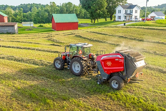 Massey Ferguson 5M tractor pulling a round baler while working in a hayfield near a farmhouse.