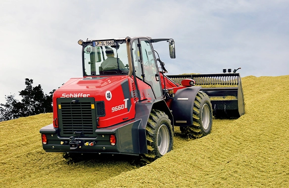 Rear view of Schäffer 9660 T telescopic loader compacting silage with a roller bucket attachment on a steep mound.