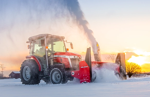 Massey Ferguson 2M Series compact tractor with snow blower attachment clearing deep snow at sunset, perfect for winter farm maintenance and snow removal operations.