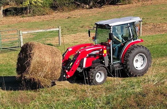 Massey Ferguson 2M Series compact tractor with enclosed cab lifting a round hay bale using a front loader, showcasing power and control for livestock feeding and hay handling.