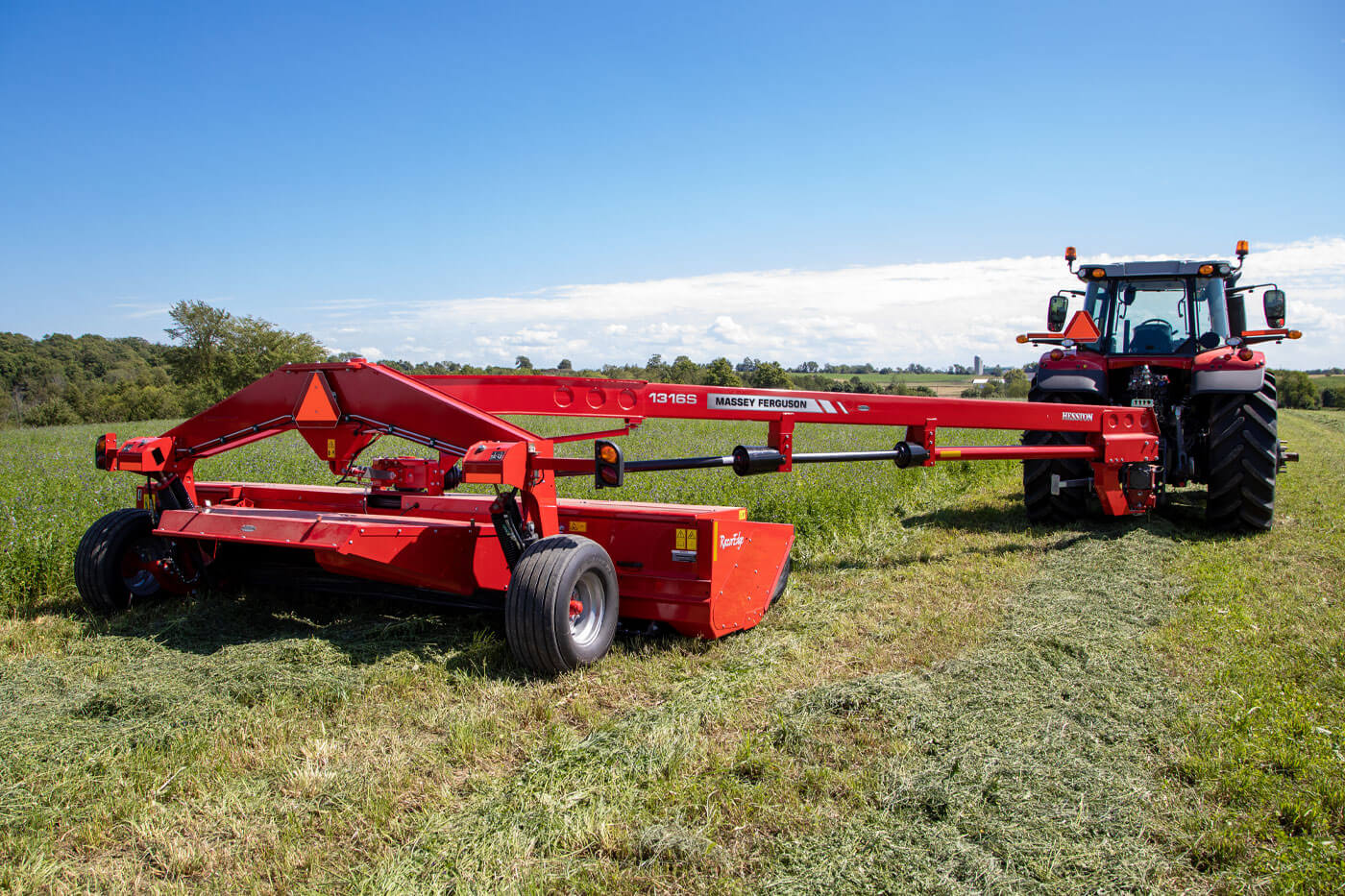 Massey Ferguson 1300 series mower conditioner in action, cutting hay in a wide field with a red MF tractor under a clear blue sky.