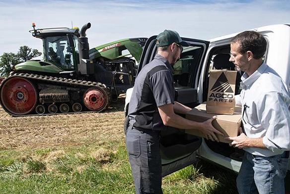 AgRevolution service technician delivering parts to a farmer in a field, with a Fendt track tractor in the background.