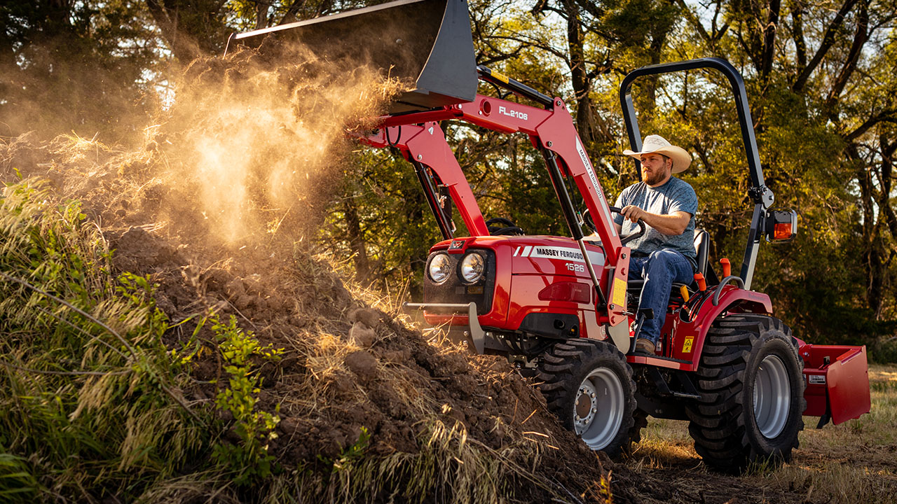 Man operating Massey Ferguson 1526 compact tractor with front loader, moving dirt on a rural property at sunset.